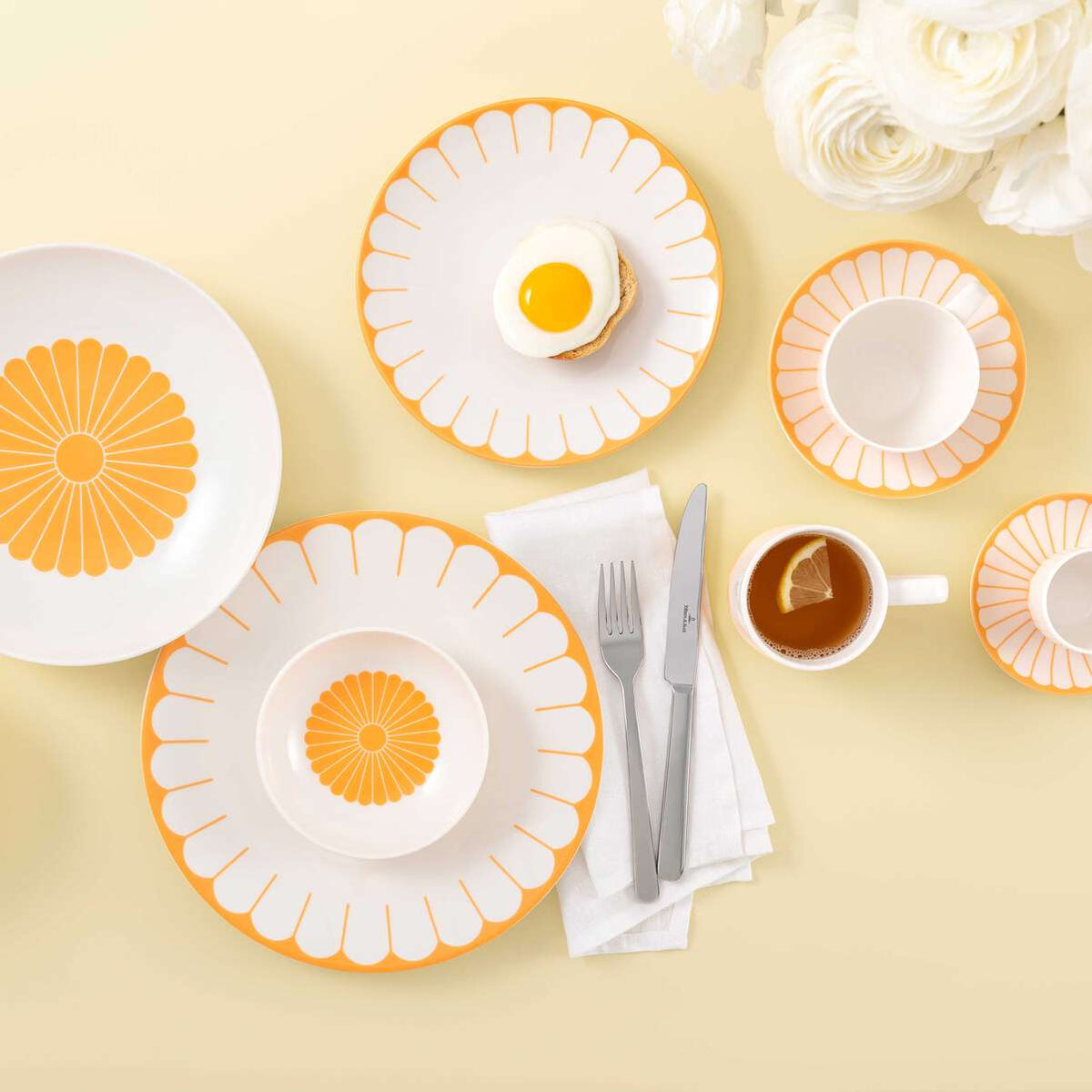 Set de table avec assiettes blanches et orange, couverts gris, une tasse de thé et une assiette avec un œuf au plat sur fond jaune.