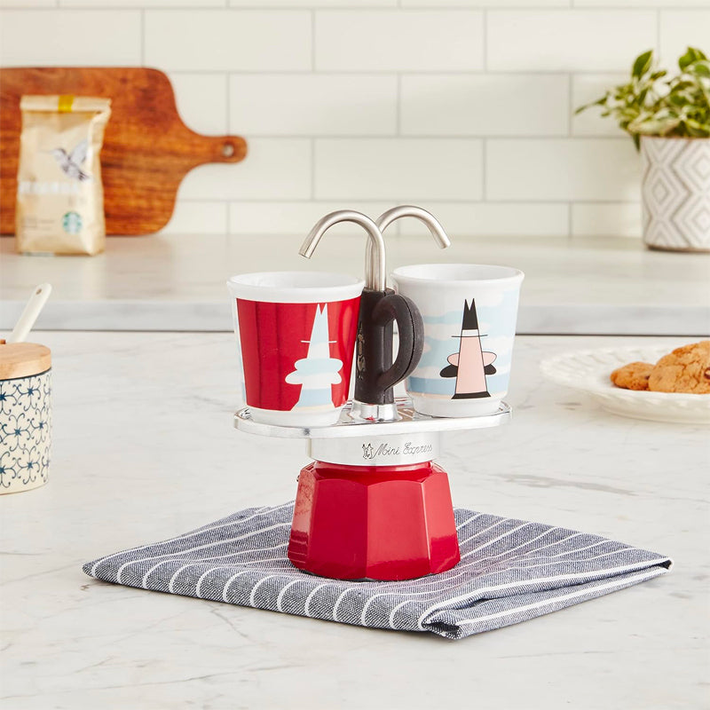 Macchina da caffè moka rossa con due tazzine decorate, appoggiata su panno a righe grigio in cucina con piano bianco e dolci sullo sfondo.