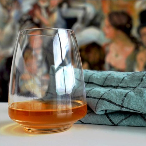 Verre transparent avec liquide orange, posé sur une surface blanche, à côté d'une serviette à carreaux bleus. Figures floues en arrière-plan.