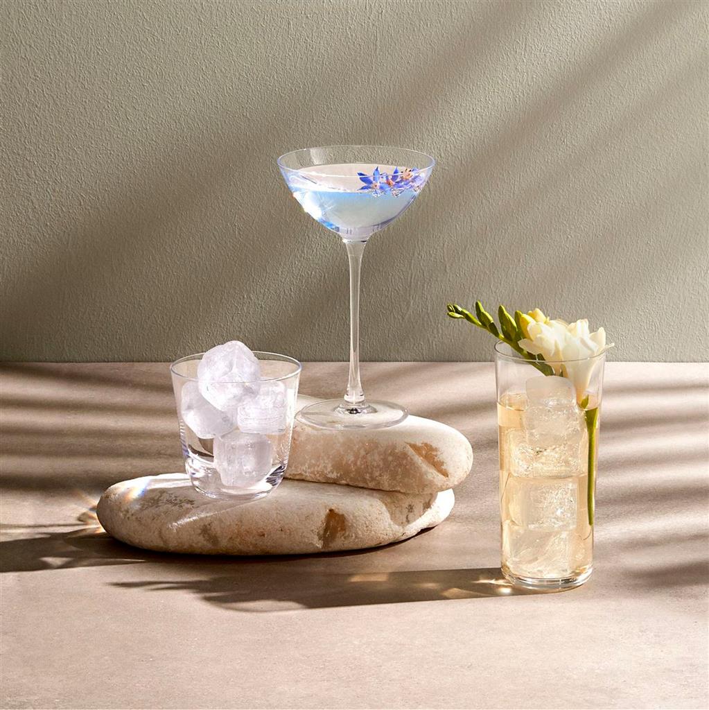 Tre bicchieri trasparenti di vetro con ghiaccio, acqua e fiore, su pietre lisce beige.