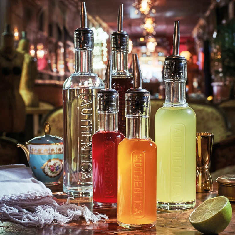 Cinq bouteilles en verre transparent avec liquides colorés rouge, orange, jaune et transparent, avec bouchons verseurs métalliques, sur table