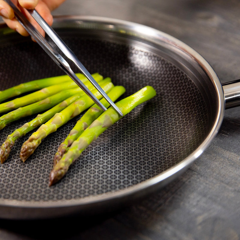 Grüner Spargel in einer schwarzen Antihaft-Pfanne mit Stahlrand und -griff, Hand mit Zange