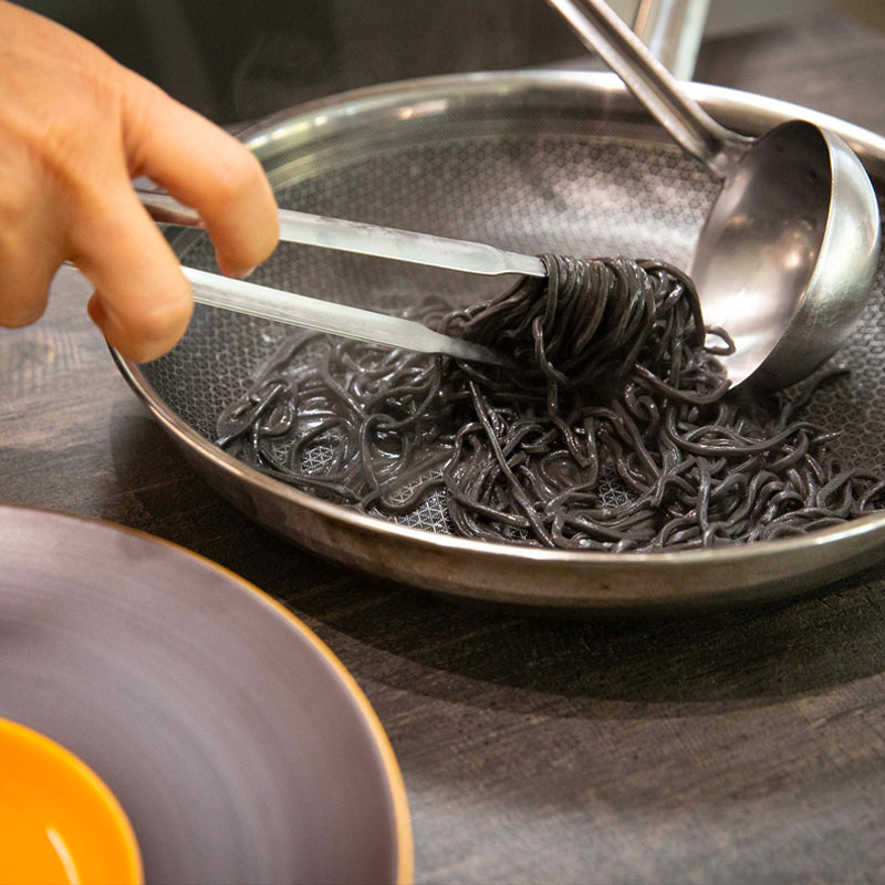 Spaghetti neri su piatto metallico con pinze in mano e mestolo di metallo, tavolo scuro in legno