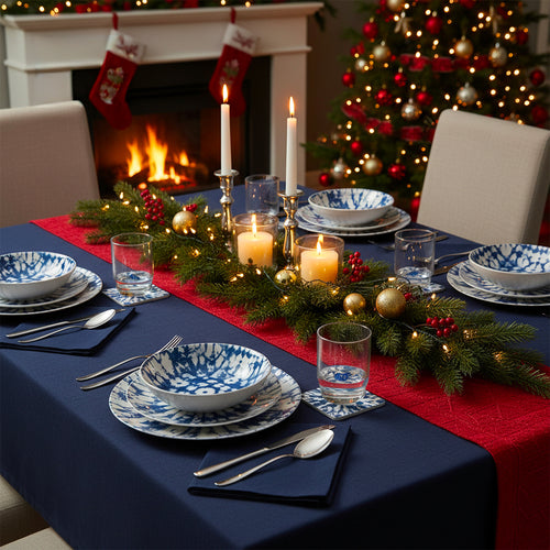 Table dressée avec nappe bleue et chemin rouge, assiettes blanches à décorations bleues, centre de table vert avec bougies et lumières, arbre et cheminée