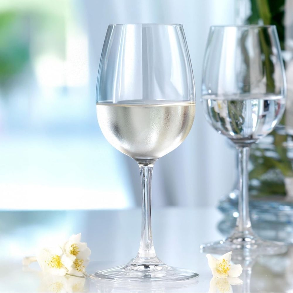 bicchiere di vetro trasparente con vino bianco, sul tavolo due fiori bianchi, sullo sfondo altri bicchieri e sfondo sfocato luminoso