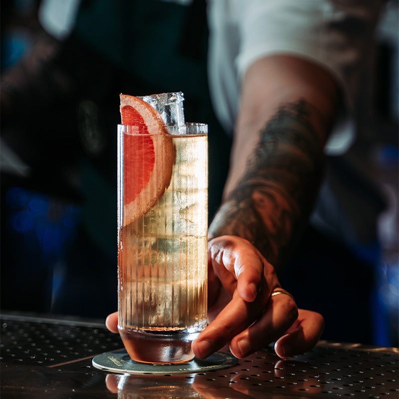 Grand verre avec boisson dorée, glace et tranche de pamplemousse rouge sur sous-verre, main tatouée au bar
