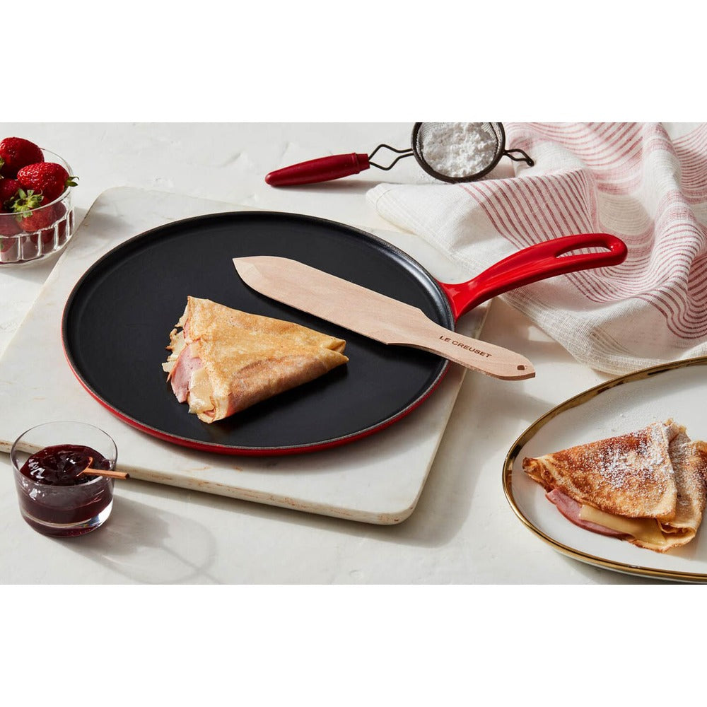 Piadina ripiena su una padella nera con manico rosso; accanto, una spatola in legno e un bicchiere di marmellata.