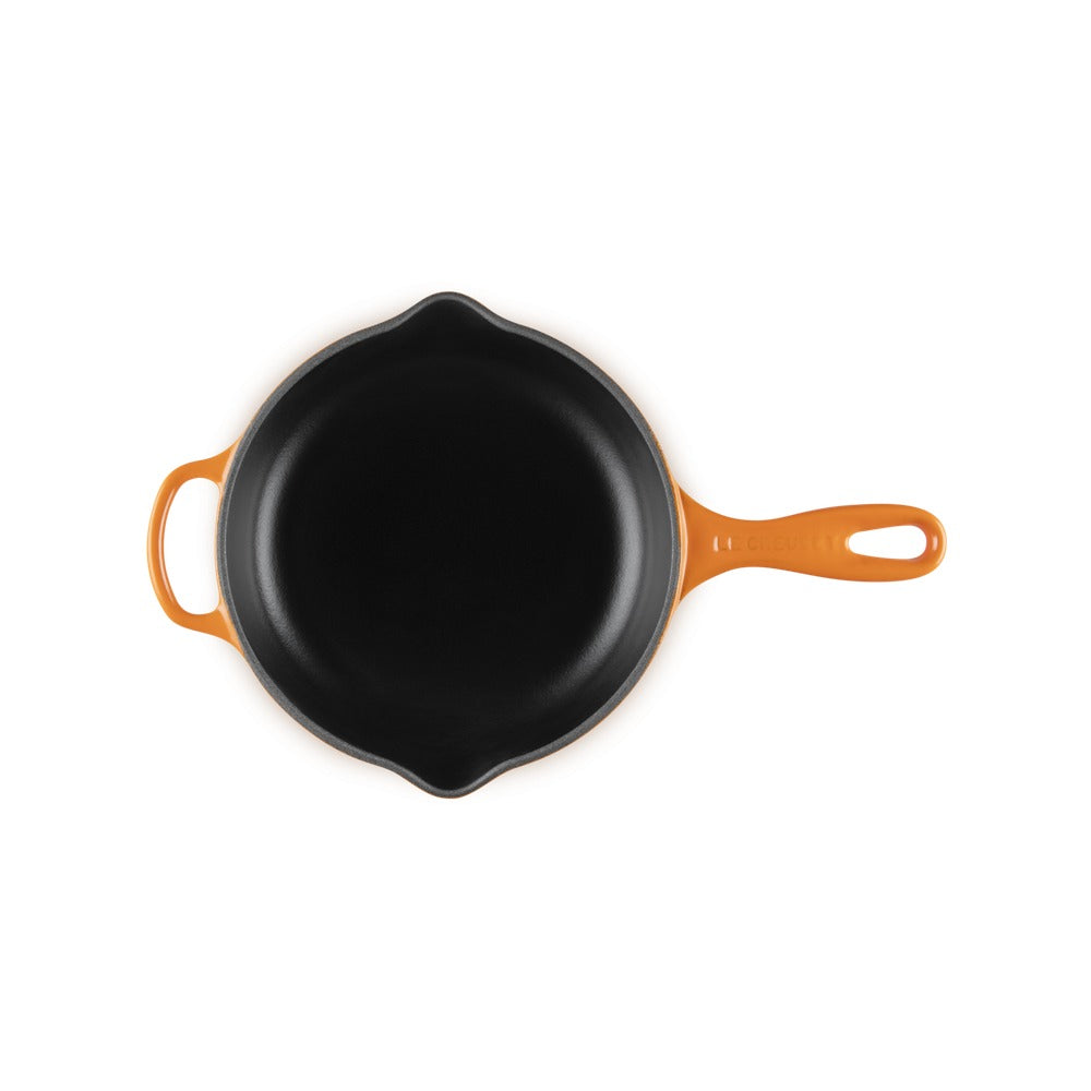 Poêle en fonte avec revêtement noir et manche orange, sur fond blanc. Forme ronde avec bords légèrement ondulés.