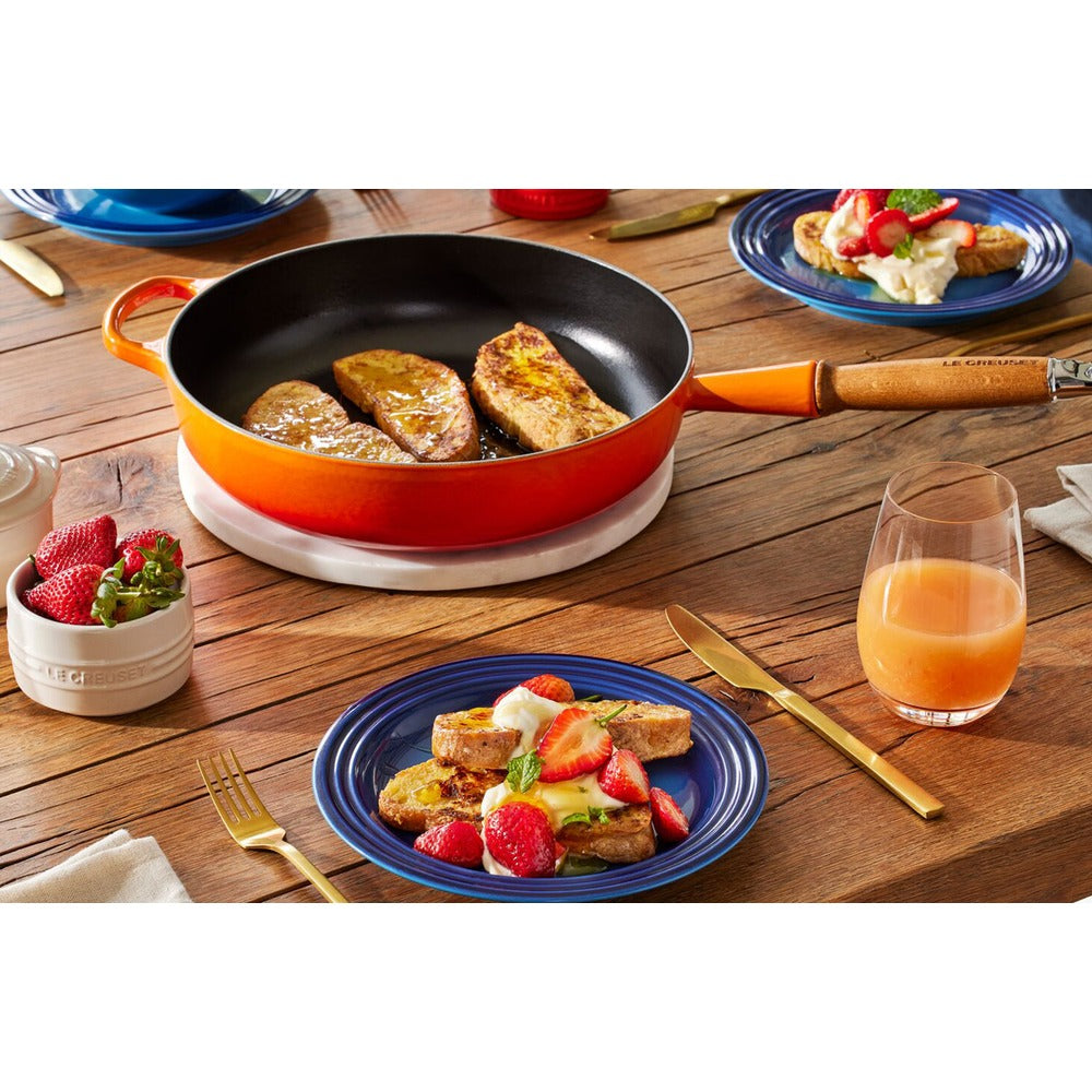 Padella arancione con fette di pane dorato, piatti blu con french toast e fragole, posate dorate e un bicchiere di succo.