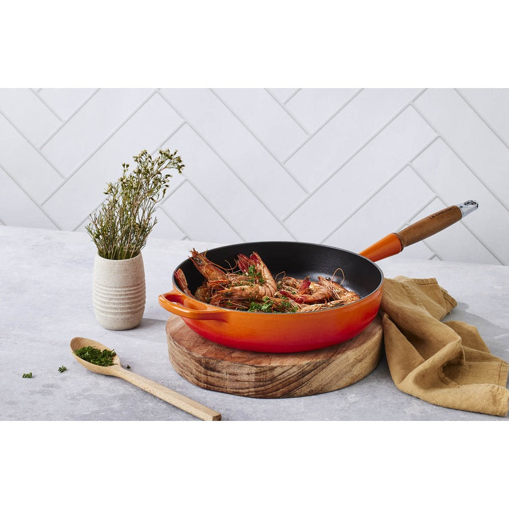 Padella rossa con gamberi, poggiata su un tagliere di legno. Accanto, un vaso con erbe e un cucchiaio di legno. Sfondo chiaro.