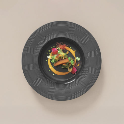 Piatto nero con bordo decorato con portata gourmet composta da salsa arancione, pezzo di pesce dorato, verdure miste e elementi decorativi verdi e