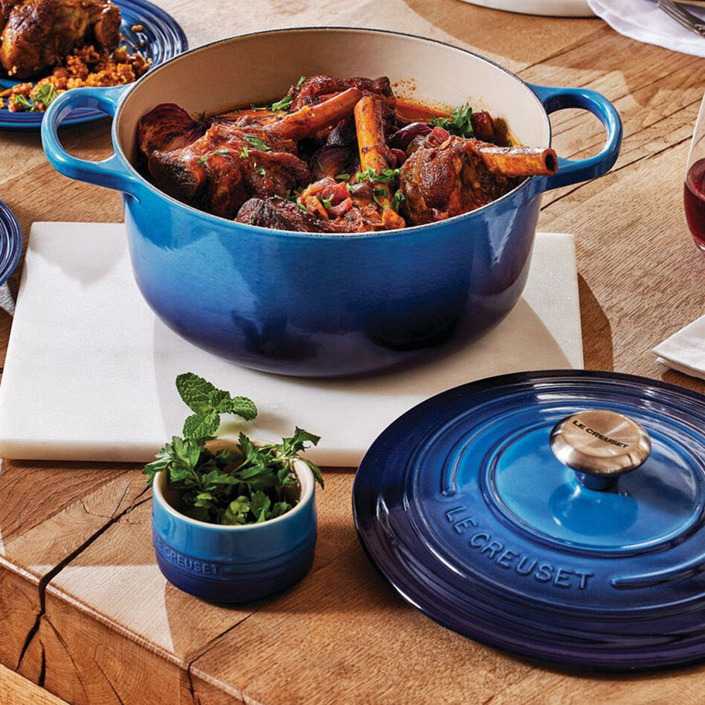 Pentola in ghisa smaltata blu con coperchio sul tavolo in legno, con spezzatino di carne e erbe aromatiche accanto a contenitore con erbe fresche.