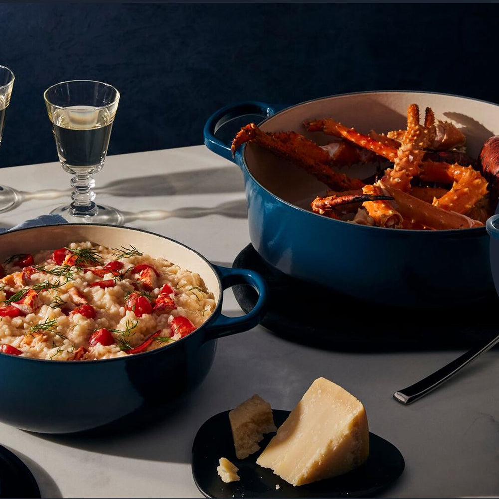 Blauwe geëmailleerde pannen met risotto met cherrytomaten en krabben, stukken kaas op zwart bord, twee glazen met transparante vloeistof op witte tafel.