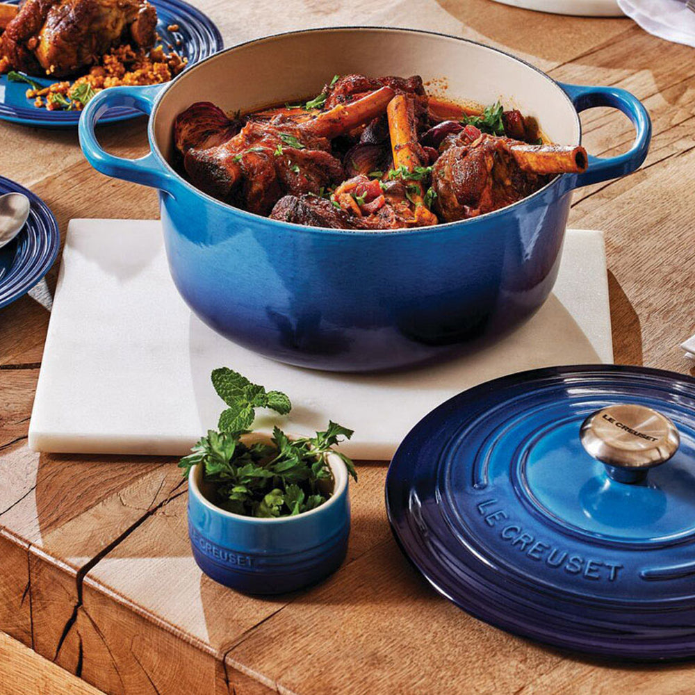 Pentola smaltata blu con brasato di carne con osso, accostata a coperchio e barattolo con erbe fresche su tavolo di legno chiaro.