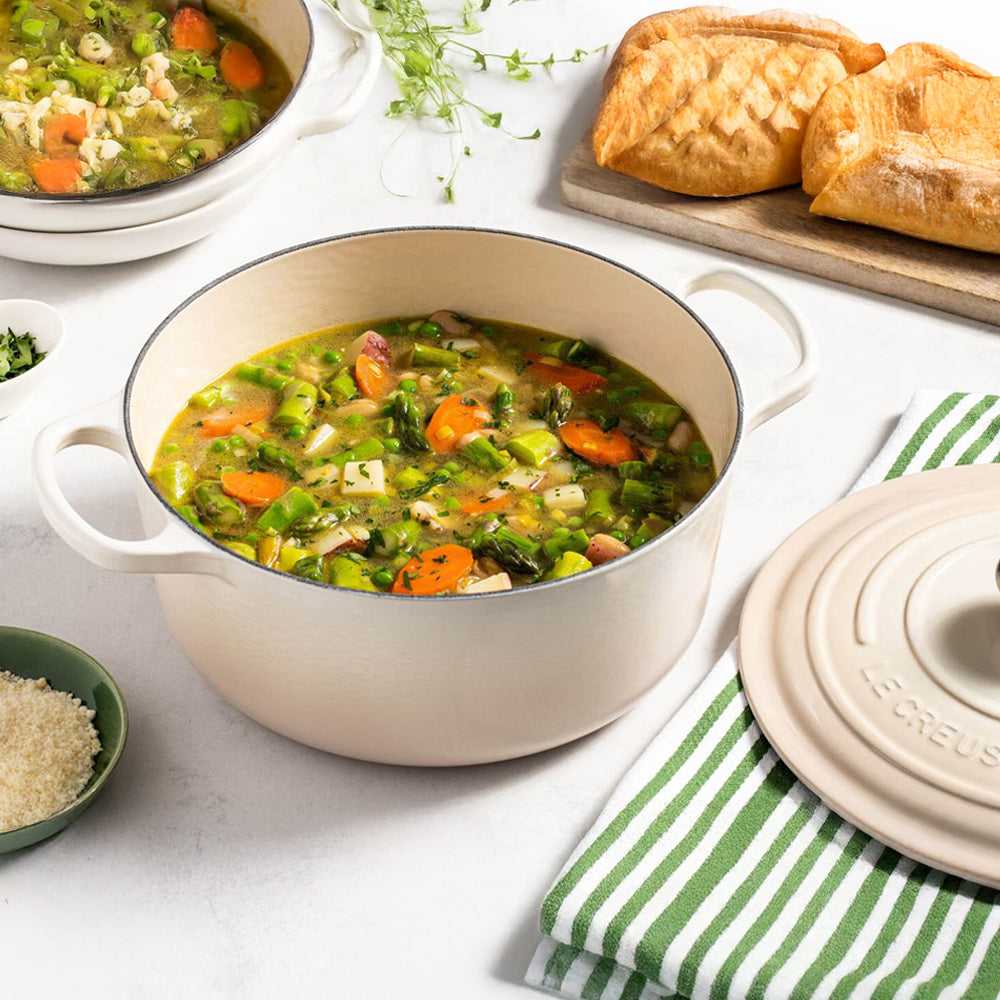 Pentola smaltata bianca con zuppa di verdure arancioni e verdi, due calzoni su tagliere di legno e asciughino a righe verdi su piano bianco.
