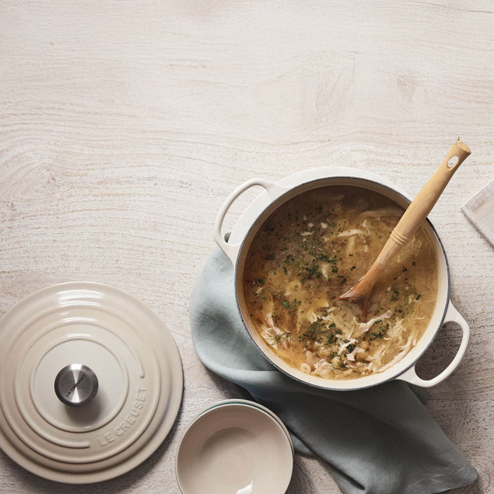 Pentola in ghisa smaltata beige con coperchio e mestolo in legno, contenente zuppa con erbe, su tovagliolo azzurro chiaro su tavolo chiaro in legno.