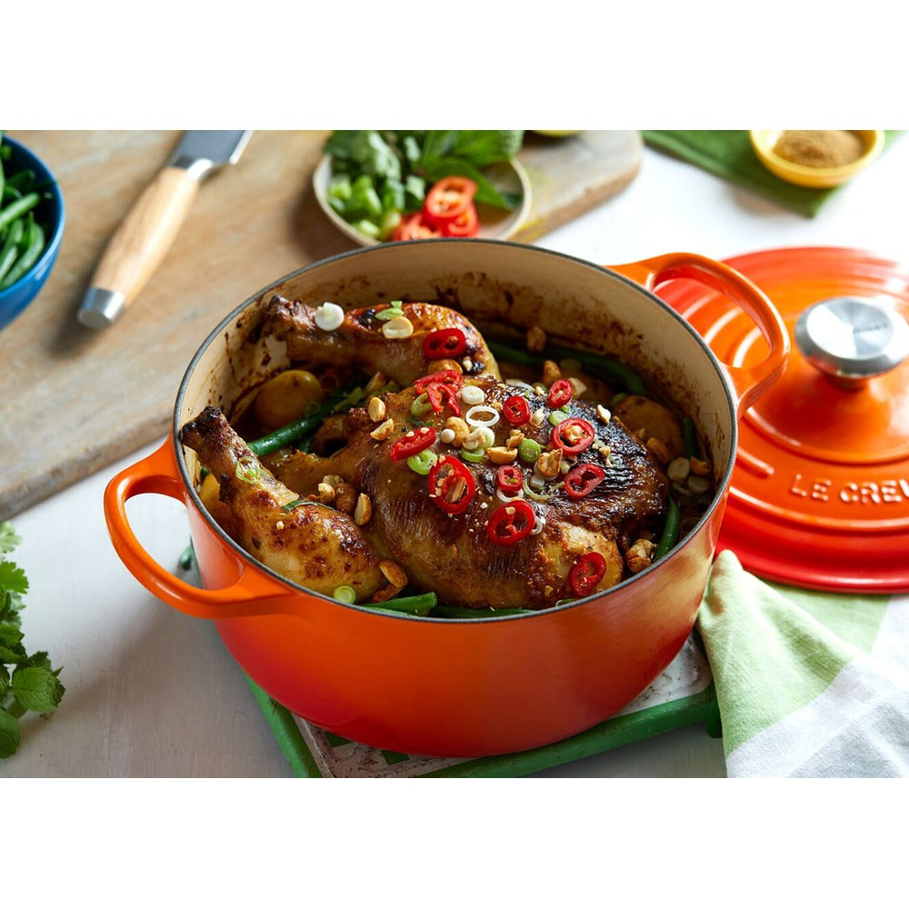 Pentola di coccio arancione con pollo arrosto guarnito da peperoncini rossi e arachidi, su un piatto verde. Sullo sfondo, verdure fresche.