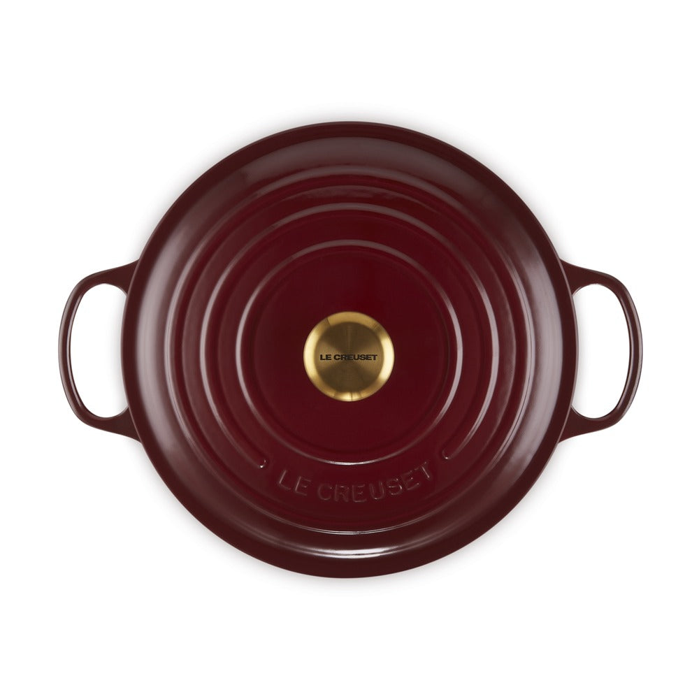 Casseruola rotonda in ghisa smaltata di colore rosso scuro con manici e logo dorato al centro.