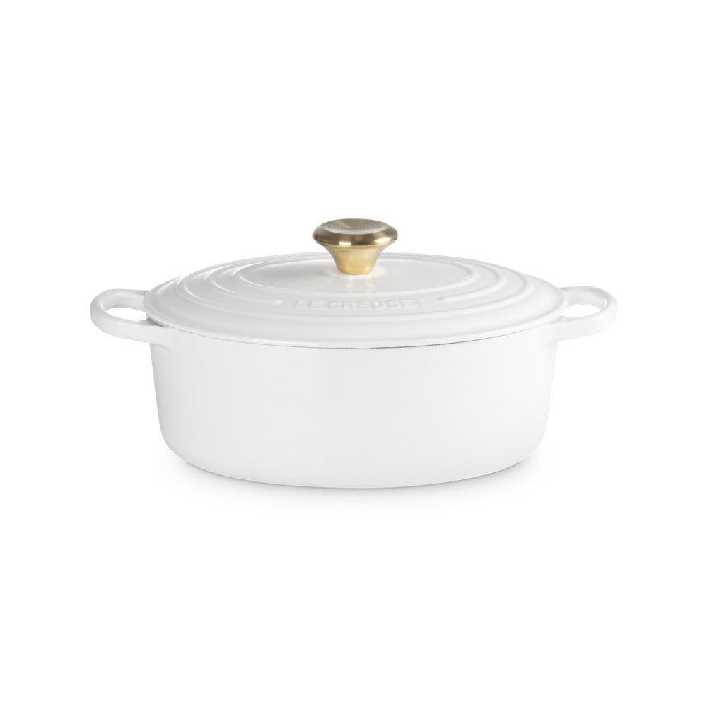 Casserole ovale blanche avec couvercle et bouton dorato, posée sur une surface neutra.