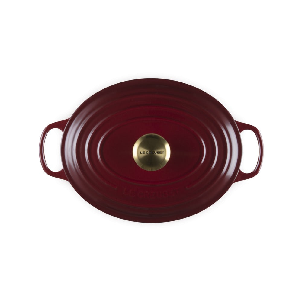 Assiette ovale rouge en fonte avec deux poignées et logo doré détaillé au centre, surface lisse et formes ondulées.