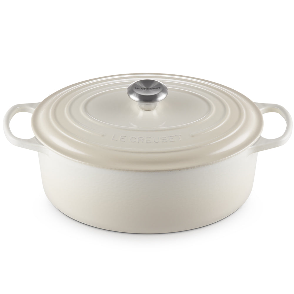 Crèmegekleurde geëmailleerde gietijzeren ovale braadpan met geïntegreerd deksel en handvatten, zilveren metalen knop met Le Creuset-opschrift, witte achtergrond.