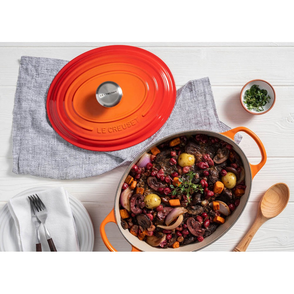 Oranje LE CREUSET gietijzeren braadpan met divers eten, bestek en servet op een lichte tafel.