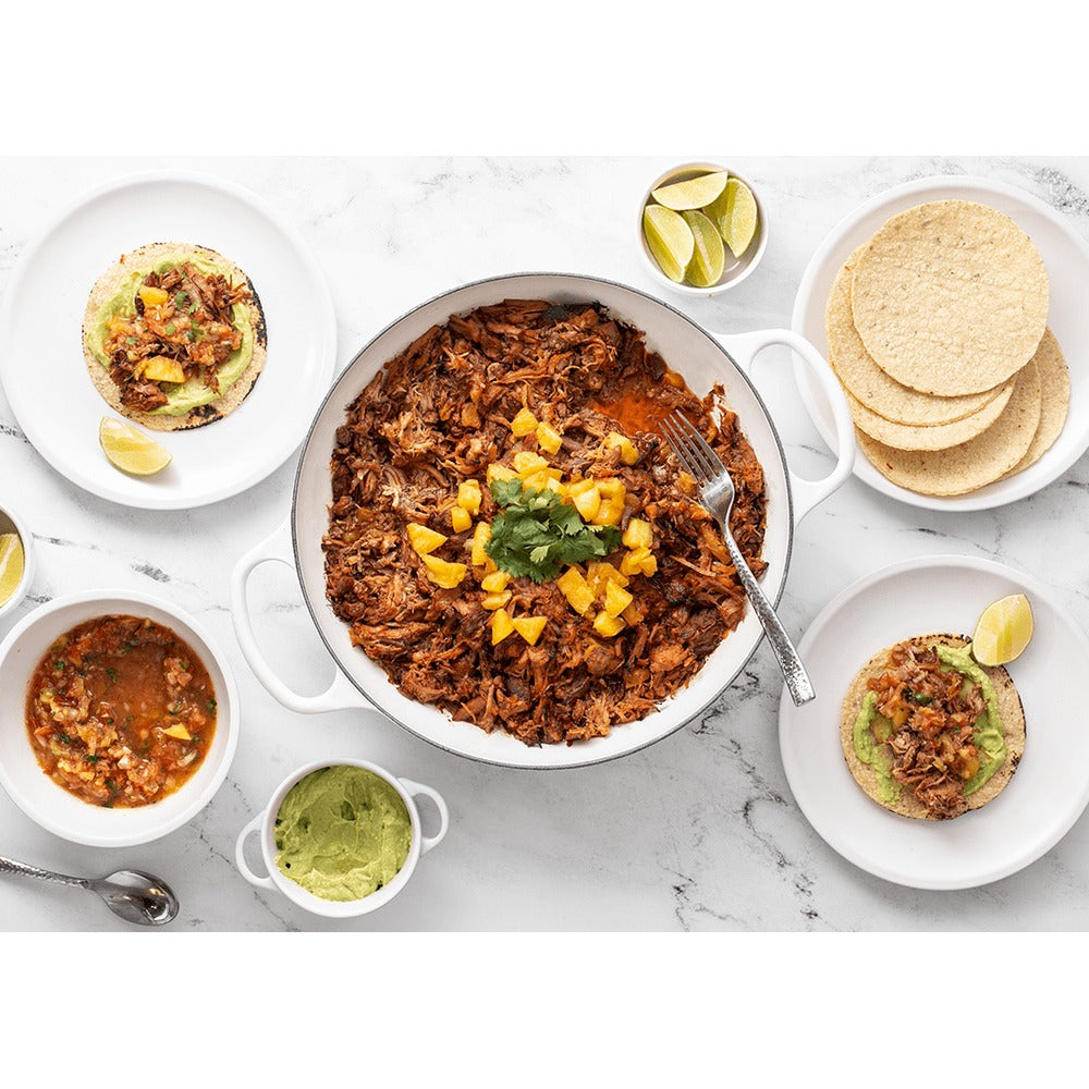 Piatto di carne sfilacciata con guarnizione di mango e coriandolo, accompagnato da tortillas, salsa e guacamole su tavolo di marmo.