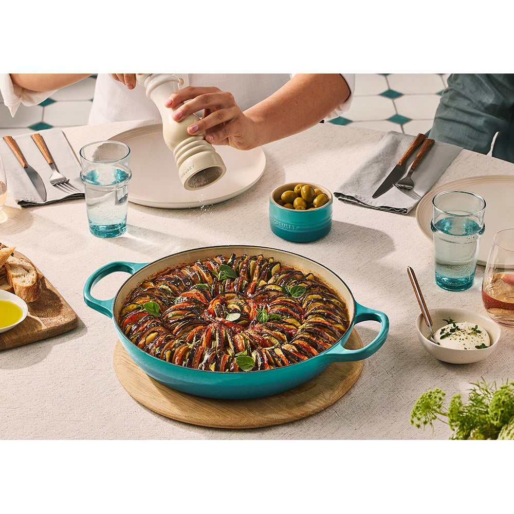 Tegame blu con ratatouille colorata al centro, bicchieri d'acqua, pane, olive, piatti bianchi e tovaglia chiara, atmosfera conviviale.