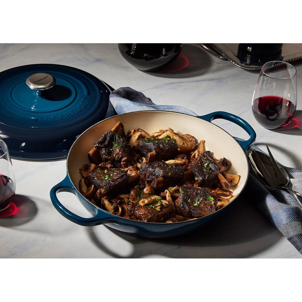 Pentola in ghisa smaltata blu con carne e funghi, guarnita con prezzemolo, e bicchiere di vino rosso sul tavolo.