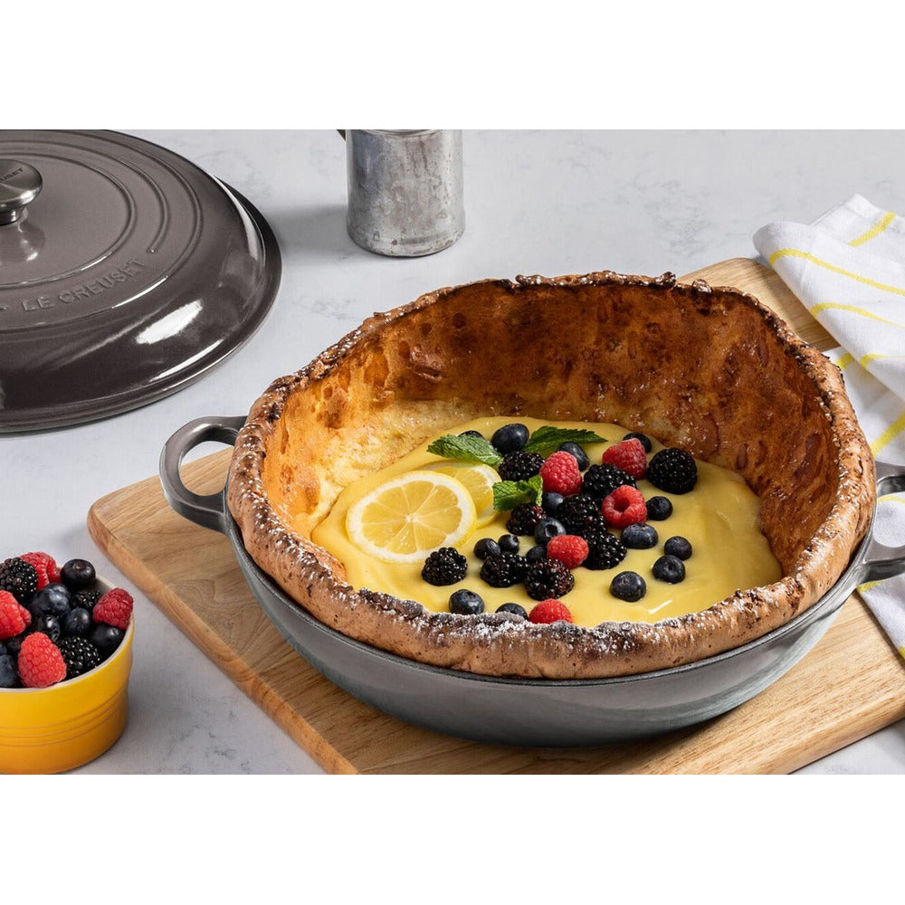Ciotola in ceramica grigia contenente un dolce con crema gialla, fette di limone e frutti di bosco. Sfondo neutro e tavola di legno.