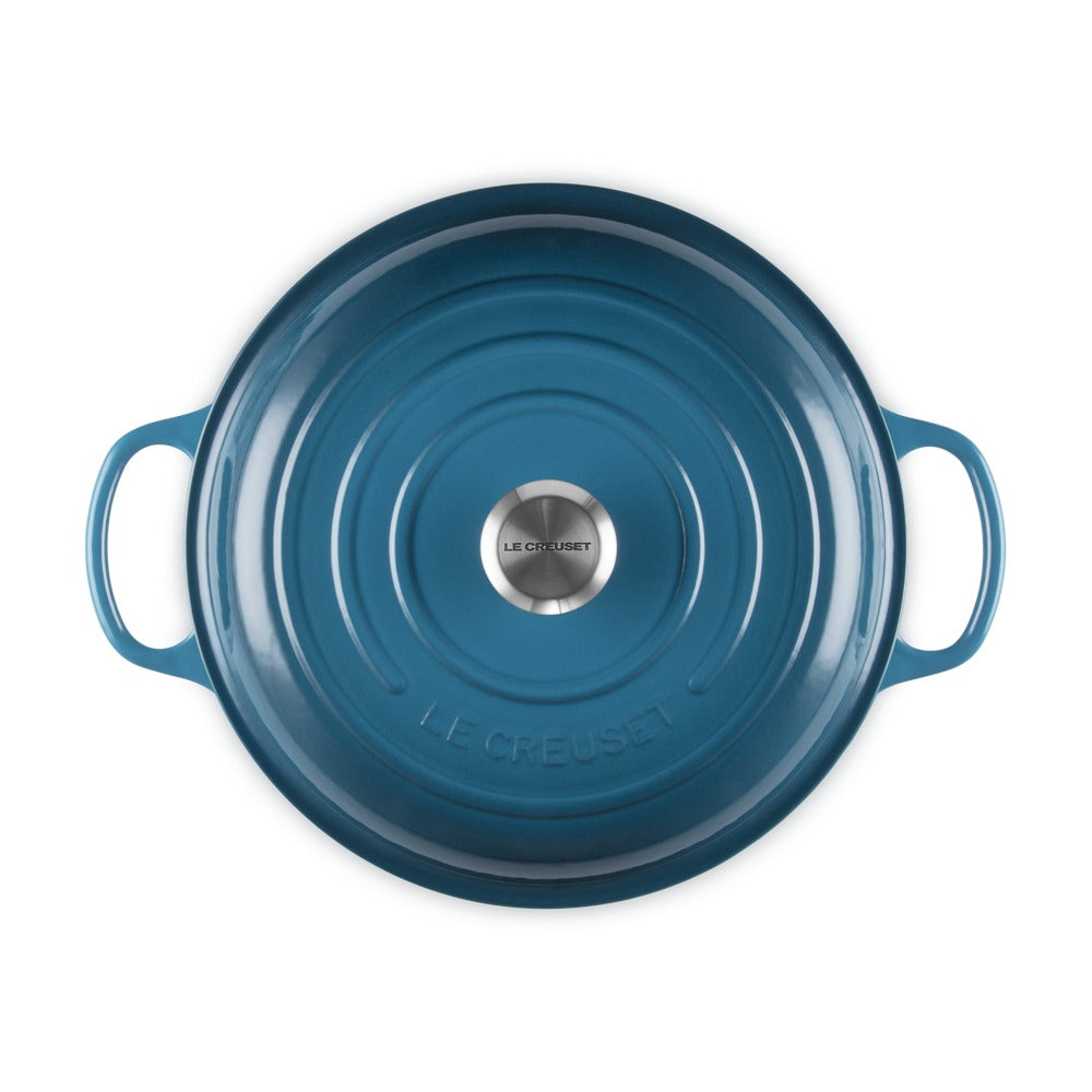 Tegame circolare in ghisa di colore blu, con manici e fondo concavo, decorato al centro con un elemento metallico.