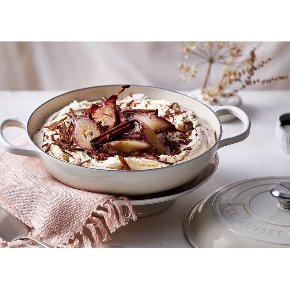 Ciotola bianca con dessert a base di panna, pere caramellate e scaglie di cioccolato, decorata con bastoncini di cannella, appoggiata su un piano