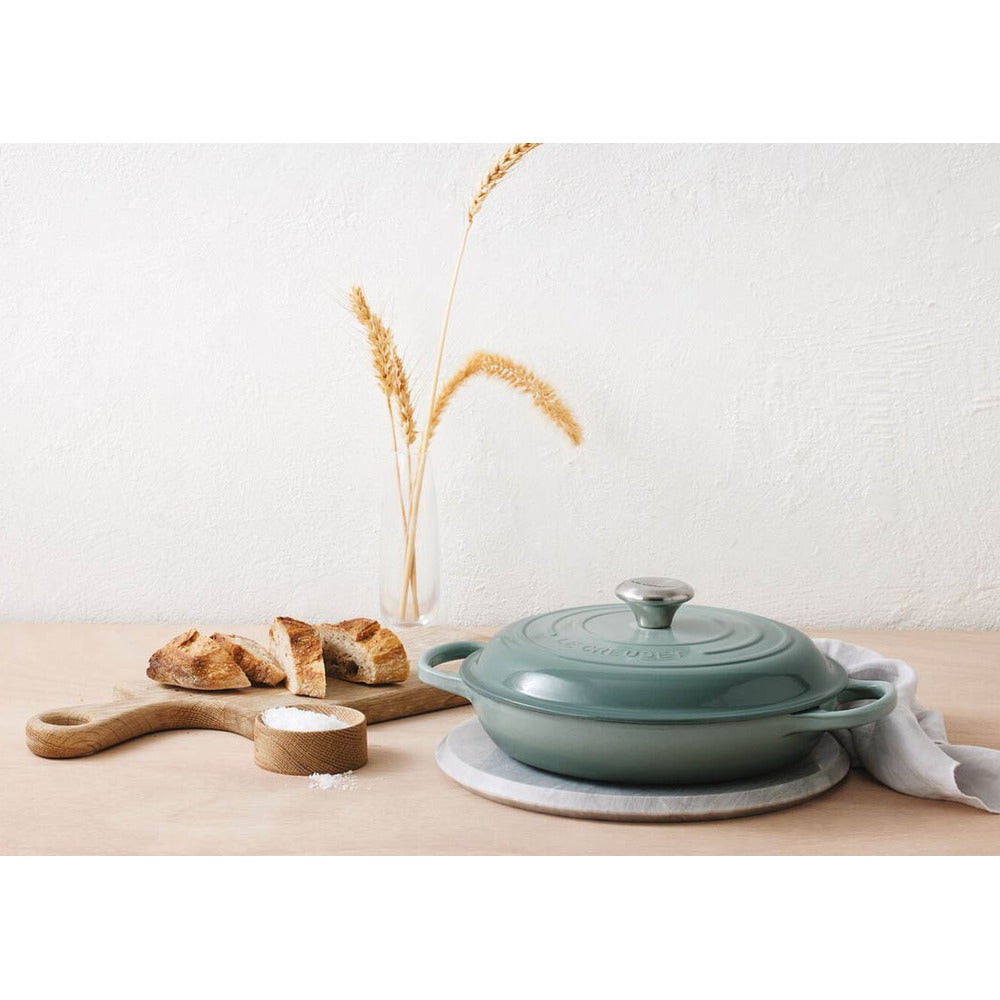 Casseruola verde in ghisa con coperchio, pane a fette su tagliere di legno, ciotola con sale e spighe d'erba secca in vaso trasparente.
