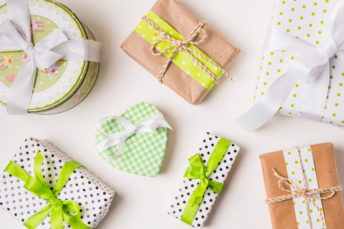 Set di pacchi regalo assortiti in carta e latta, con fiocchi verdi, bianchi e spago su sfondo chiaro.