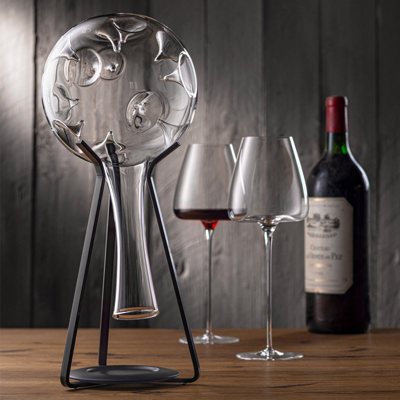 transparenter Glasdekanter auf schwarzem Ständer mit zwei Weingläsern und Flasche auf Holztisch