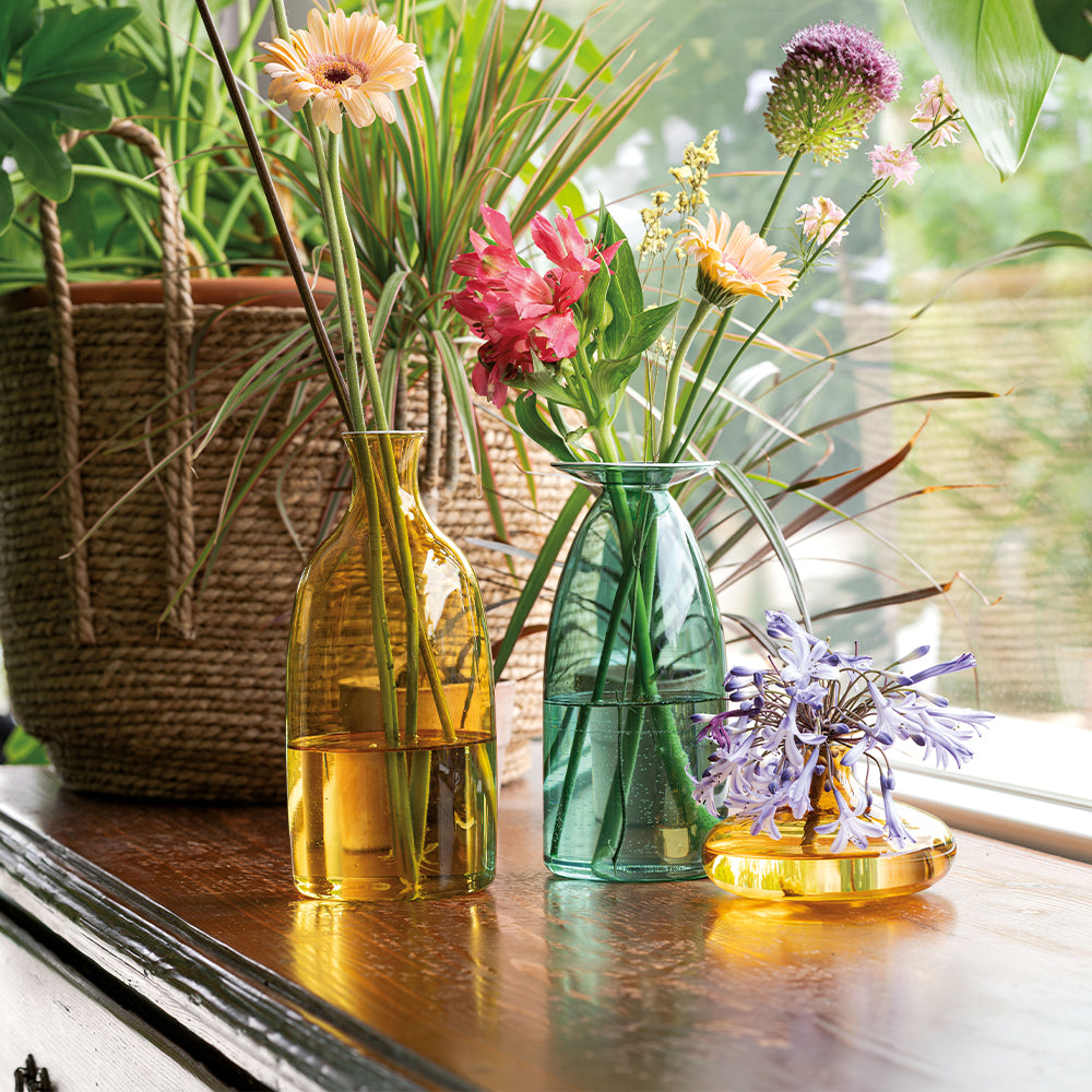 Gekleurde glazen vazen in geel en groen met verse bloemen, op houten tafel, in lichte omgeving versierd met planten.