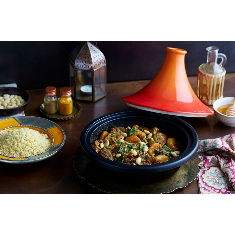 Un tajine nero con carne e frutta secca, riso basmati in un piatto, spezie in barattoli e lanterna decorativa sullo sfondo.