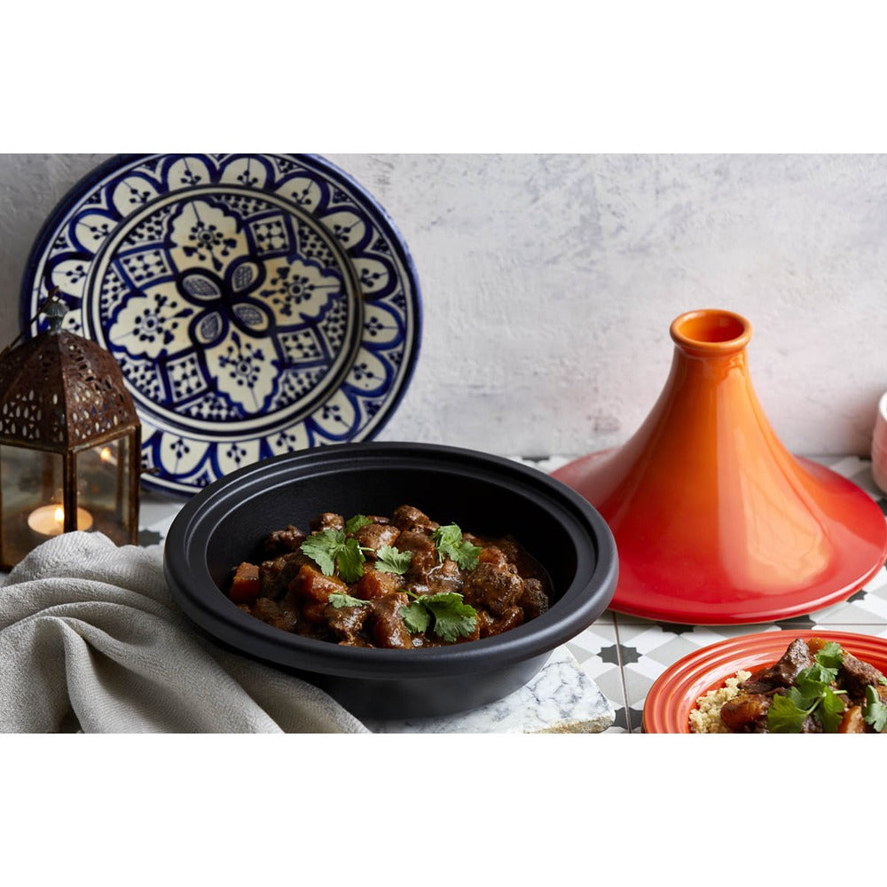 Piatto di stufato in una ciotola nera, decorato con coriandolo, accanto a un tajine arancione e un piatto rosso. Sfondo in piastrelle.