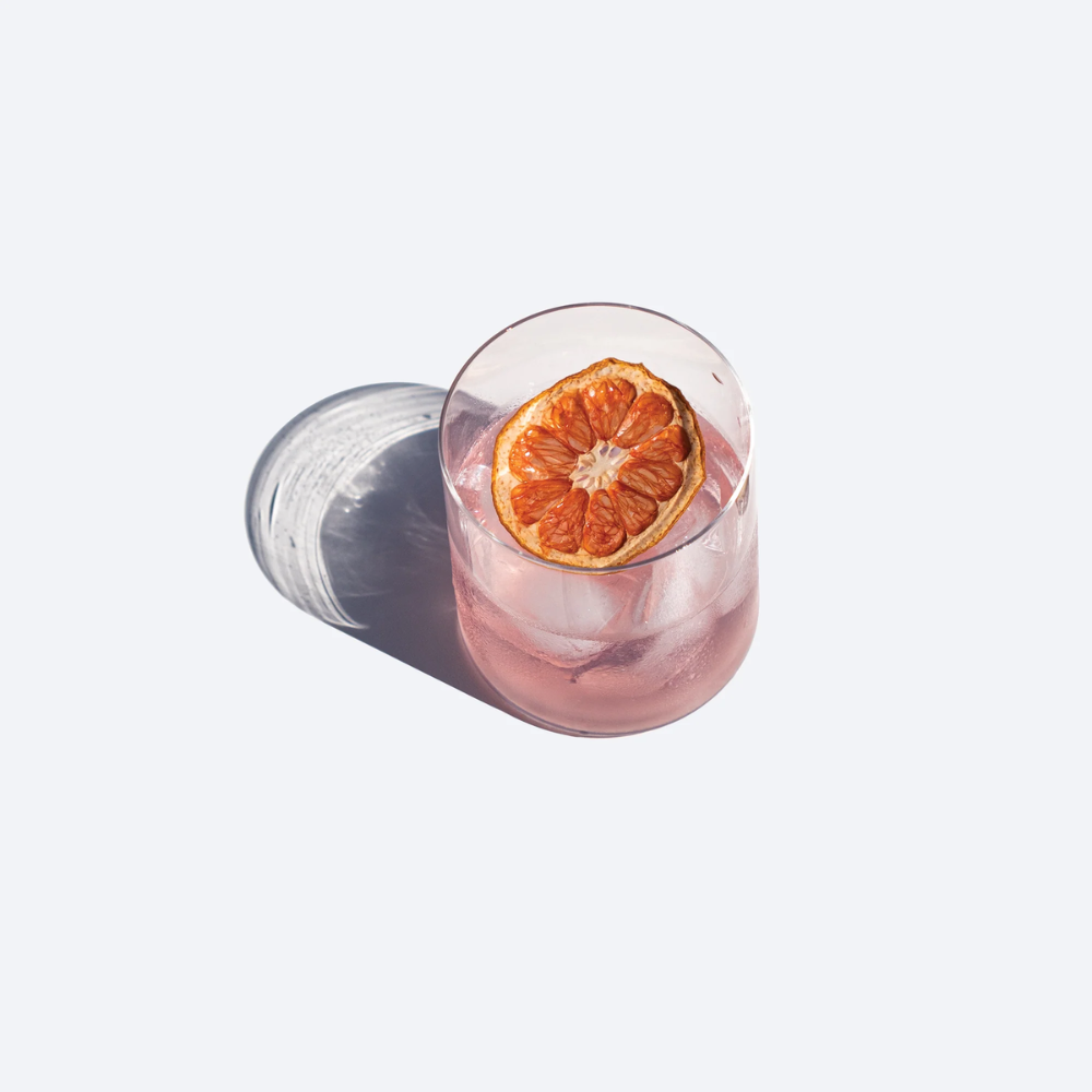 bicchiere trasparente con bevanda rosa, cubetti di ghiaccio e fetta di agrume essiccato, su sfondo chiaro con ombra netta