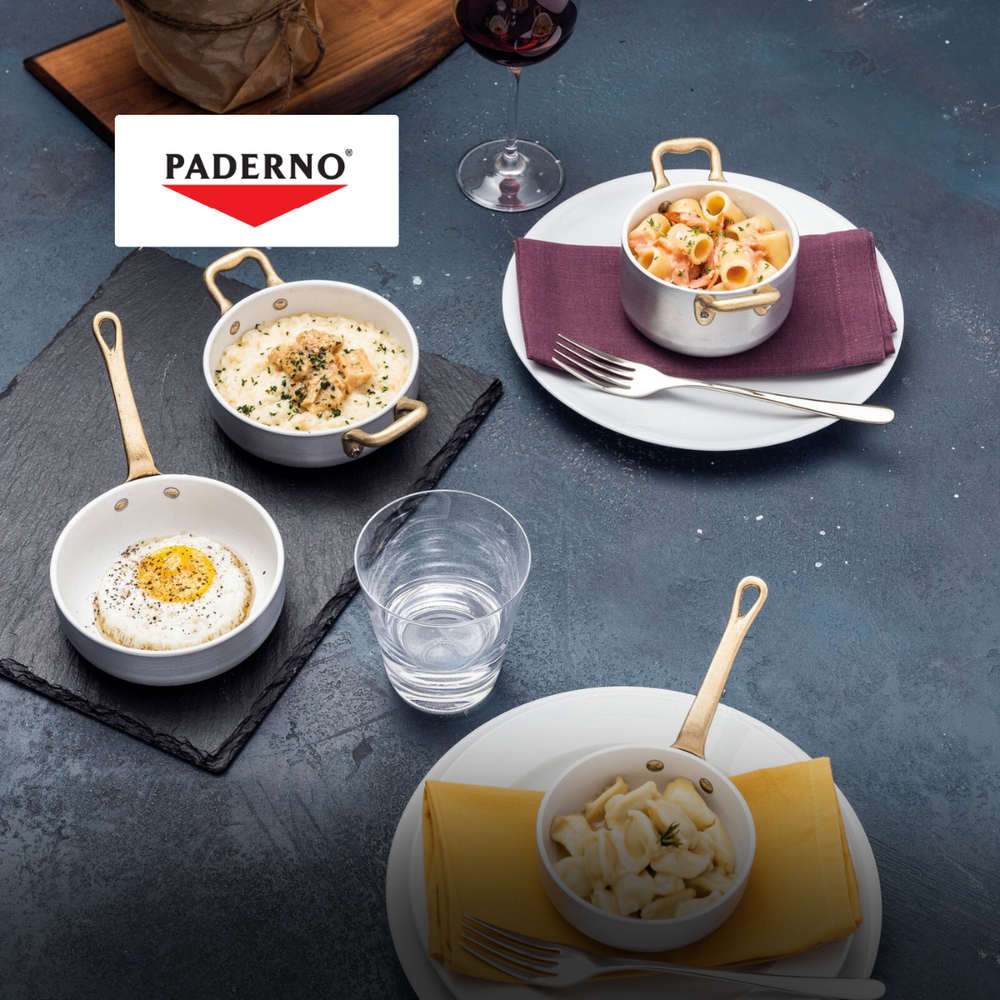 Pentole Paderno in alluminio con manici dorati contenenti cibo e un bicchiere d'acqua su tovaglioli colorati