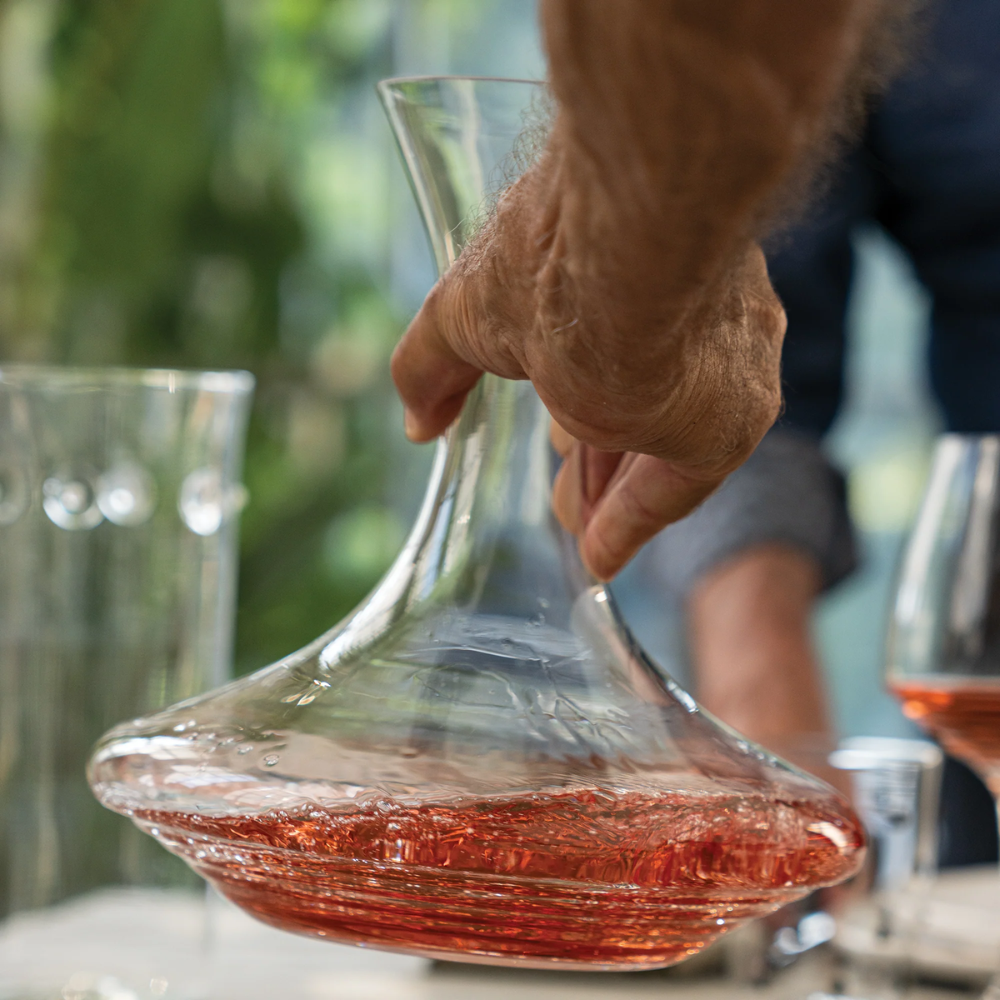 mano che tiene una caraffa di vetro trasparente con vino rosato, sullo sfondo vetri e figura sfocata di persona in ambiente interno luminoso