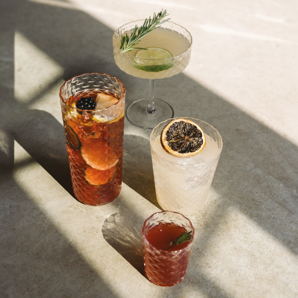 Cuatro vasos de vidrio facetado con cócteles coloridos, decorados con rodajas de fruta y ramitas, sobre superficie de cemento claro