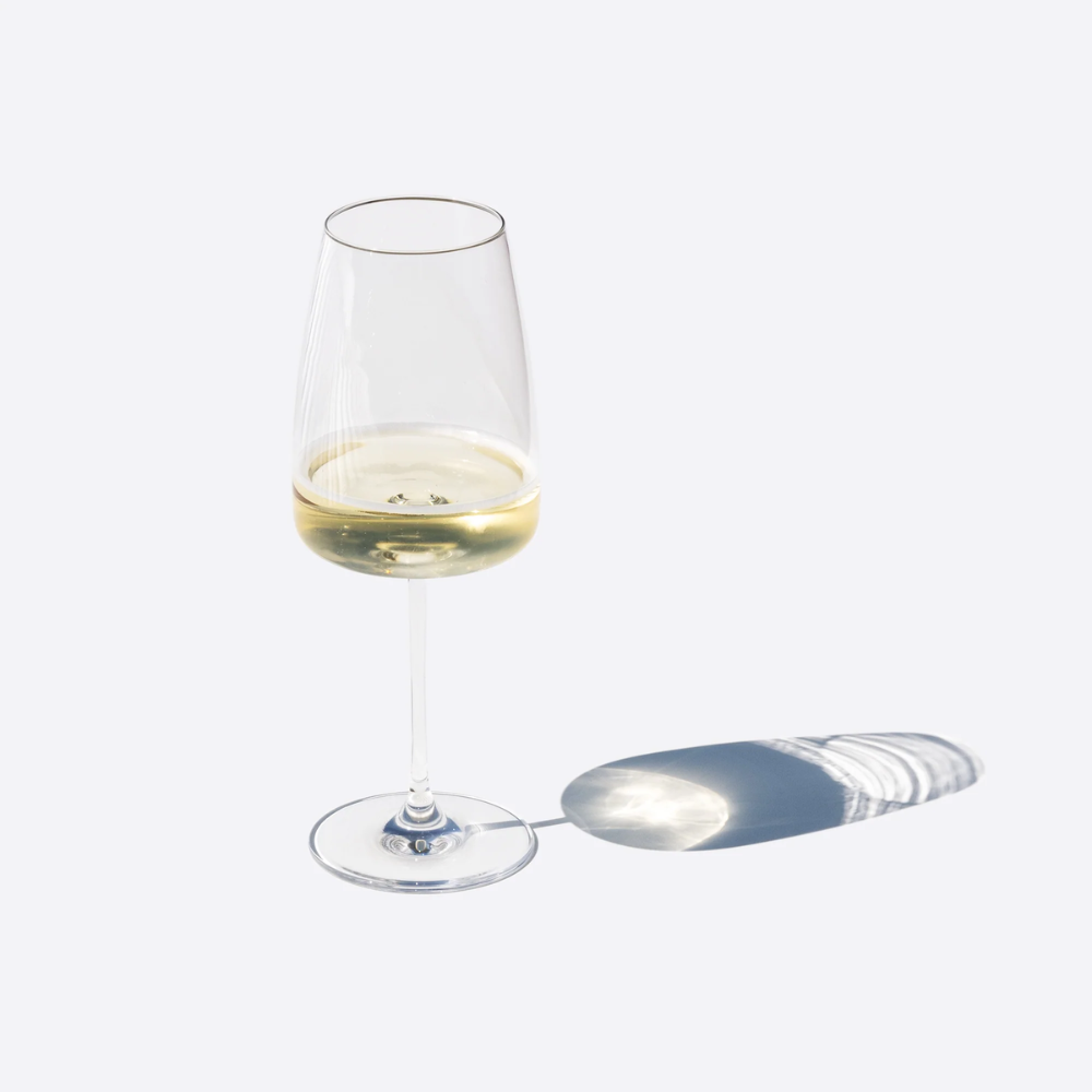 Bicchiere di vetro trasparente con vino bianco, forma conica e base piatta, ombra su sfondo chiaro.