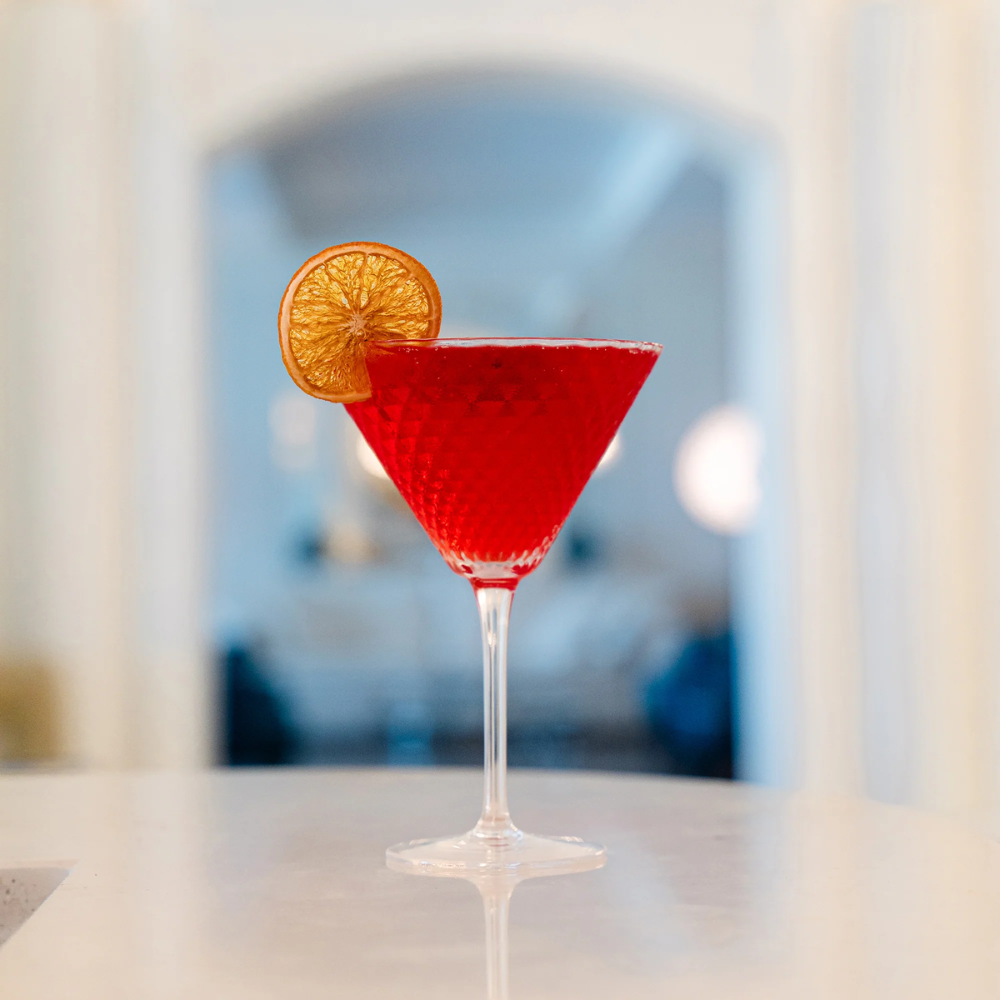 cocktail rosso in bicchiere di vetro inciso con fetta di agrume essiccato, su superficie bianca con sfondo sfocato interno casa