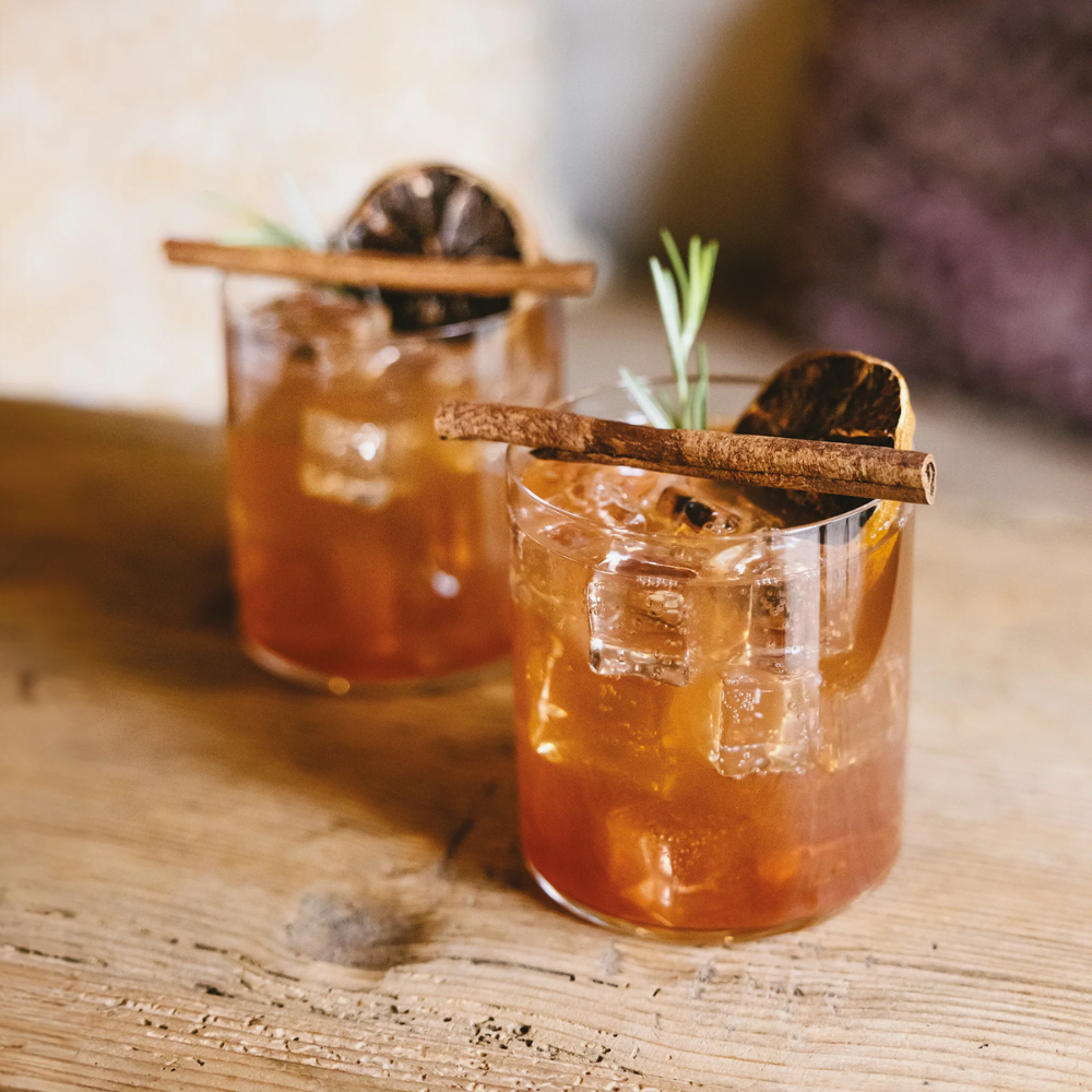 Due bicchieri di vetro trasparente con cocktail ambrati, ghiaccio, stecca di cannella, fetta di agrume essiccato e rametto verde su tavolo