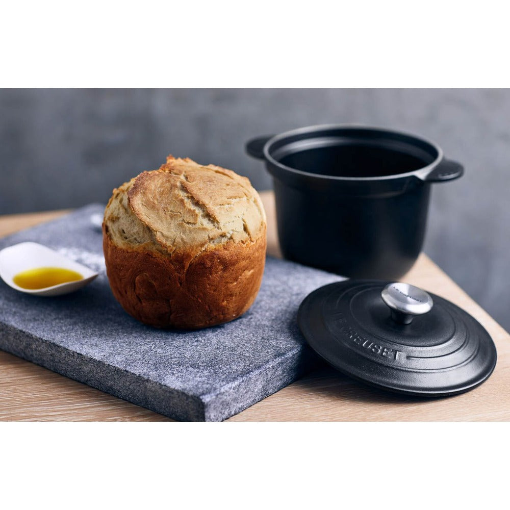 Pain doré sur planche en pierre grise, à côté d'une casserole noire et d'une assiette avec huile d'olive.