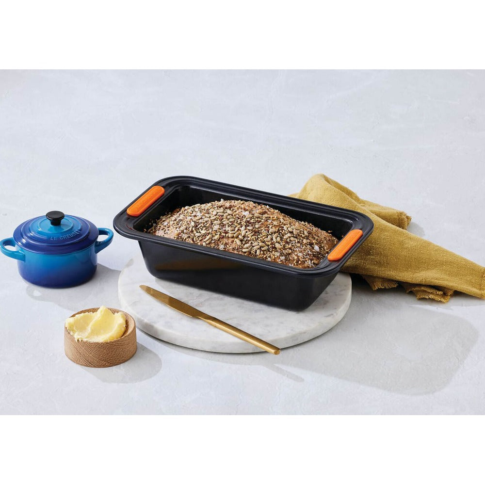 Conteneur noir pour pain avec pâte et noisettes, serviette jaune, couteau doré, petite casserole bleue et beurre sur planche.