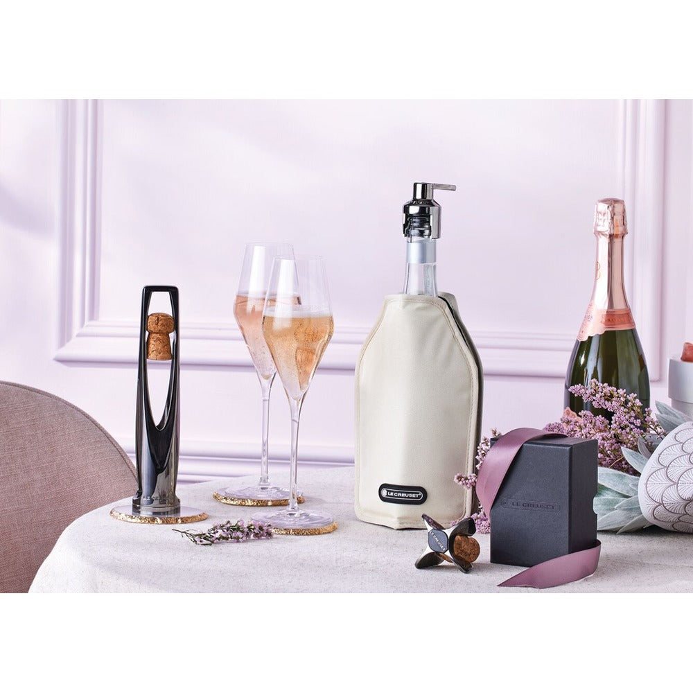 Due flûte di champagne su un tavolo, una bottiglia con copertura beige e un cavatappi nero, in un ambiente elegante con decorazioni floreali.