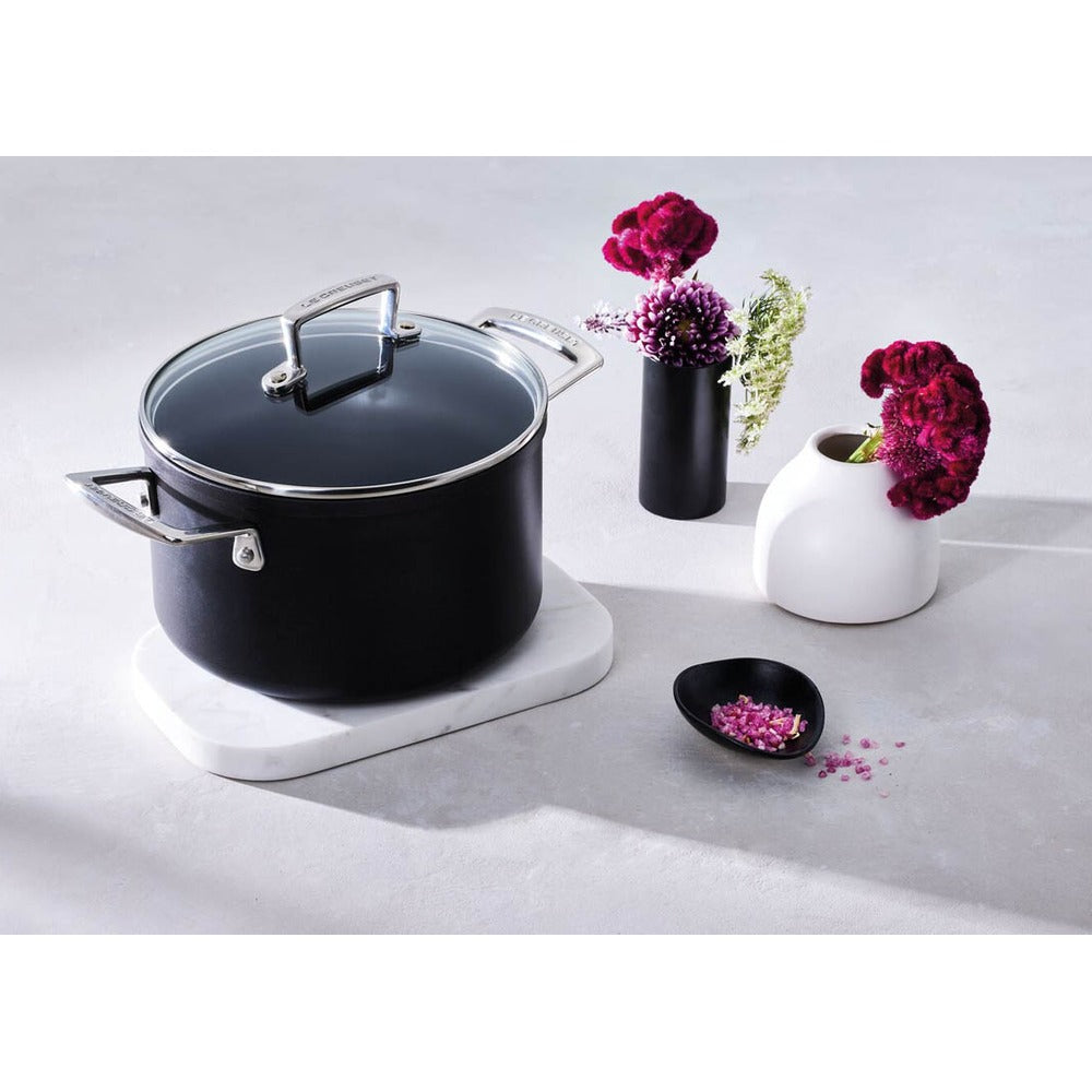 Olla negra con tapa transparente sobre salvamanteles de mármol, junto a jarrones blancos y negros con flores moradas.
