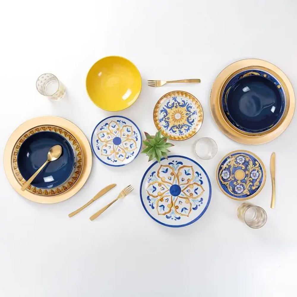 Set di piatti colorati su un tavolo bianco: ciotola gialla, piatti blu con decorazioni e posate dorate, un vaso con pianta.